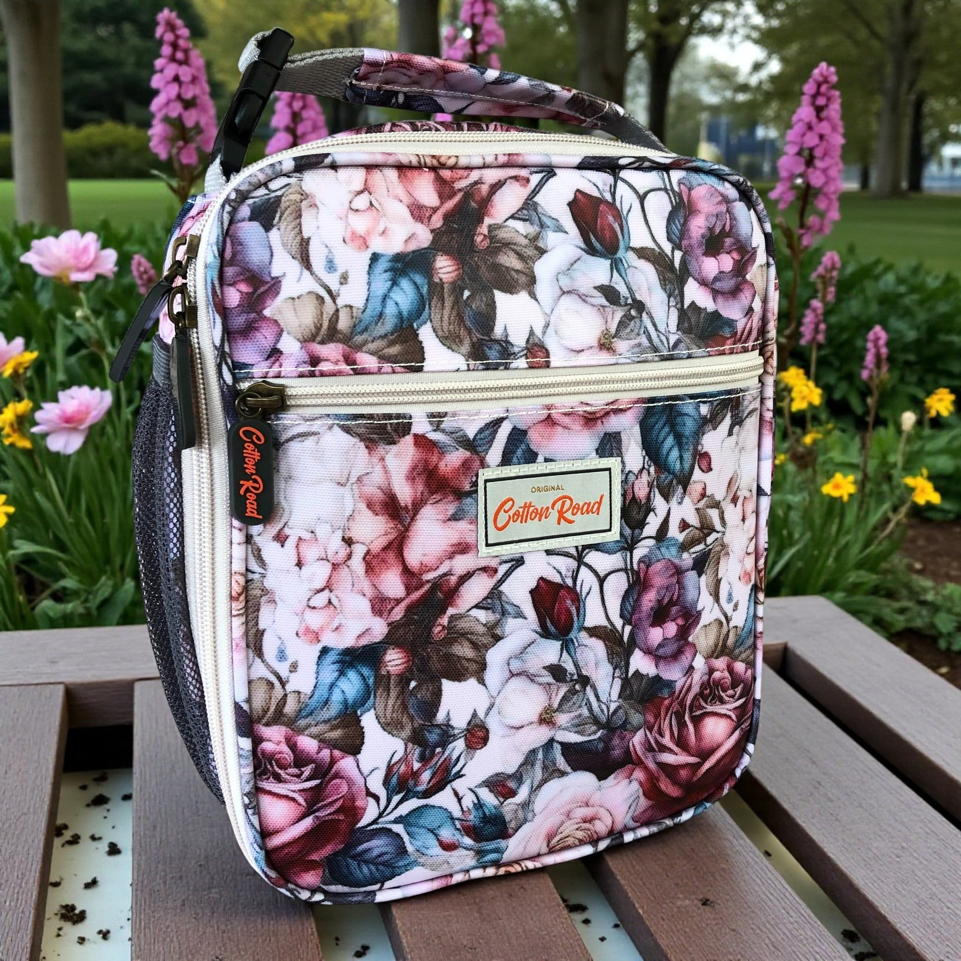 COTTON ROAD COOLER PINK Floral-patterned lunch bag on a wooden bench with flowers in the background