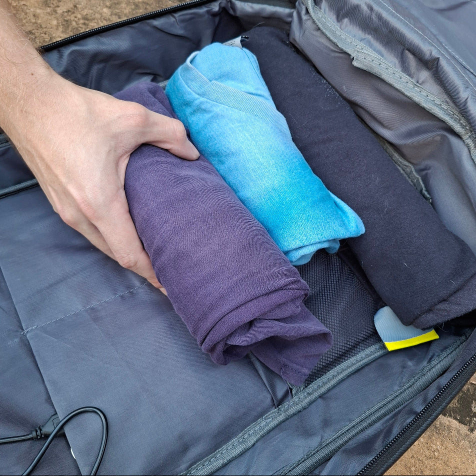 Person packing rolled-up clothes into an open Vivace blue medium size backpack with USB port visible