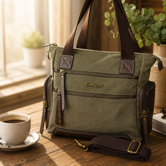 Cotton Road green canvas shoulder handbag with brown leather trim styled on a wooden table in a warm lifestyle setting with coffee cup, books and indoor plants. Available from We Sell Bags South Africa